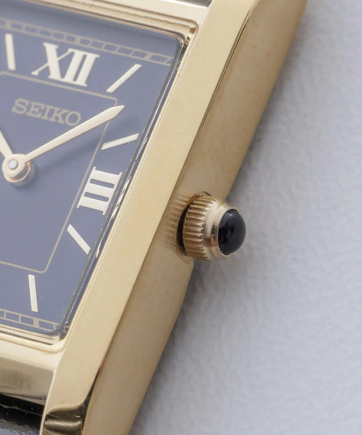 Close-up of a Seiko rectangular watch with a gold-tone case, blue dial, Roman numeral hour markers, and a black cabochon-style crown accent on a light textured background.